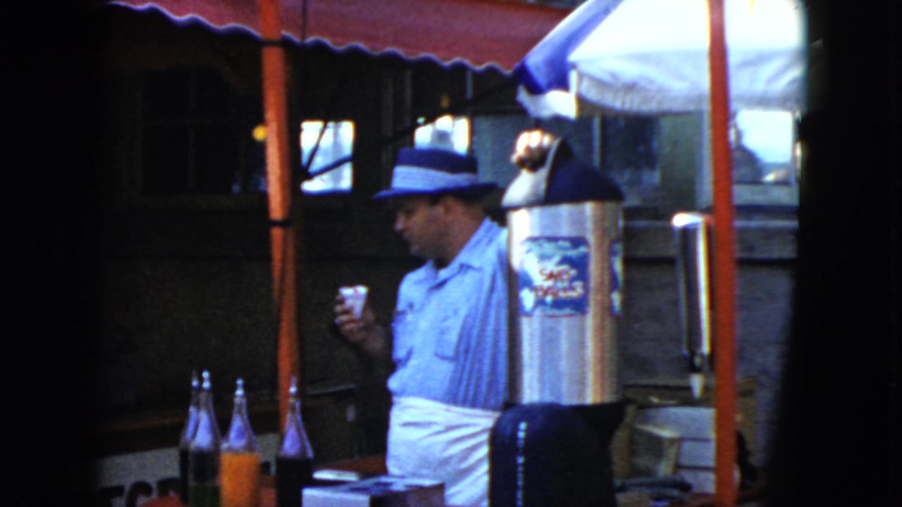 1951 Cleveland home-movie frame showing a man in a fedora and white apron holding a cup near bottles and a metal dispenser at a stall.