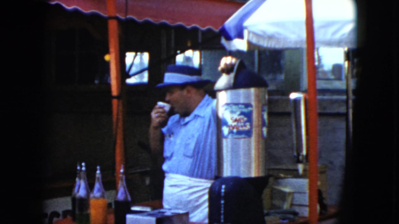 1951 Cleveland frame of a man in apron and fedora sipping from a cup at a food or drink stall with bottles nearby.