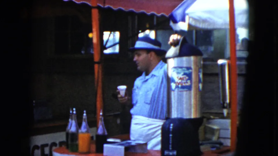 1951 Cleveland home-movie still showing the man holding his cup lower while standing near colorful bottles and a metal drink dispenser.