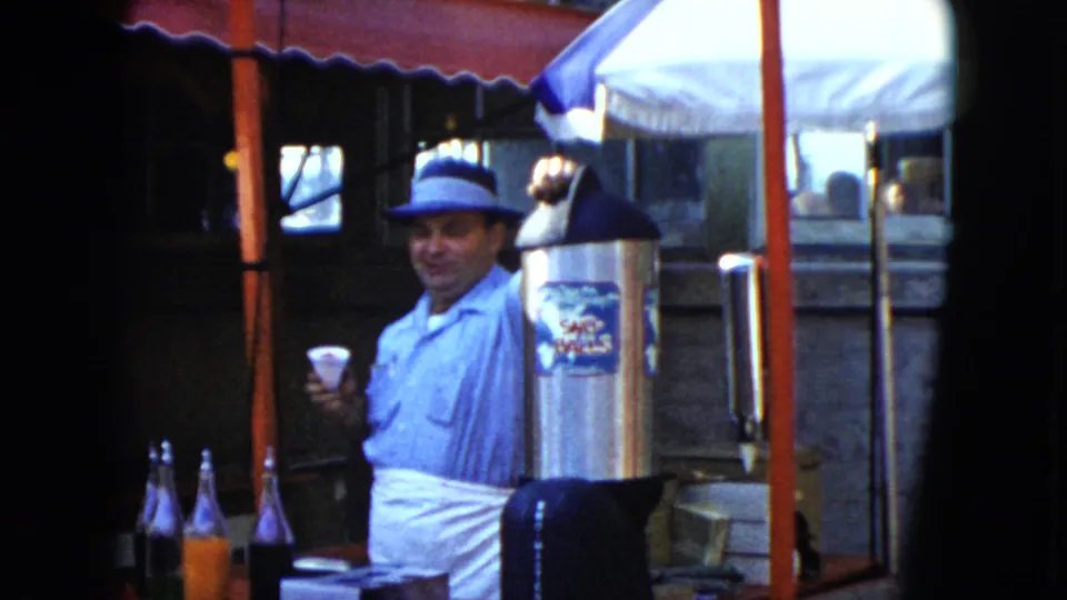 1951 Cleveland frame with the man facing the camera, cup in hand, beside a labeled metal dispenser under a red awning.