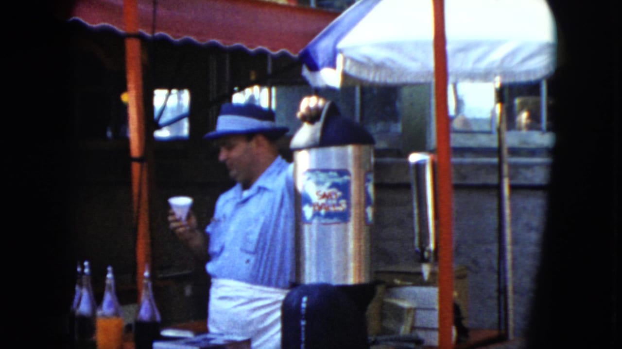 In 1951 Cleveland home-movie footage, a man stands at a sidewalk refreshment stall beside a silver dispenser and bottled drinks.