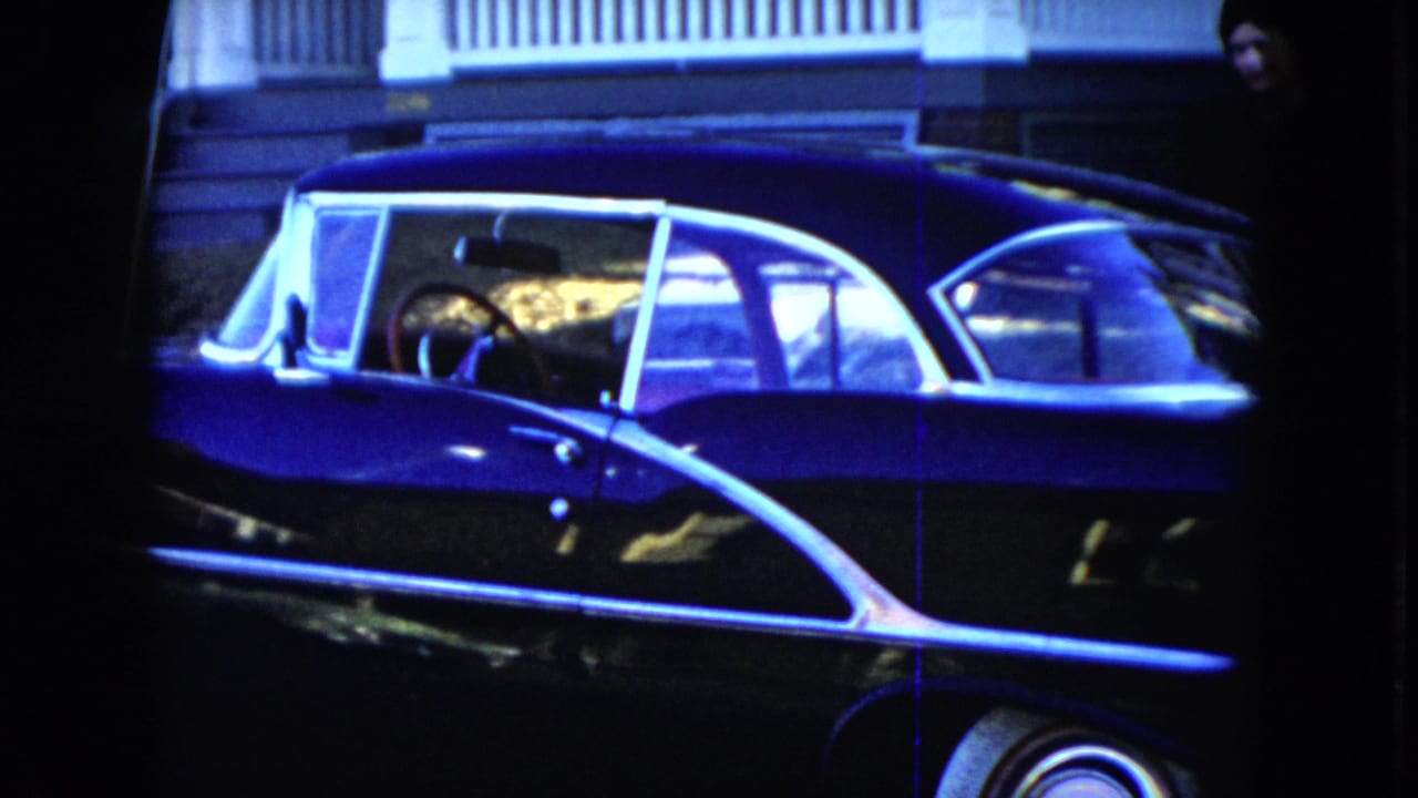 Dark blue mid-century sedan parked beside a house porch.
