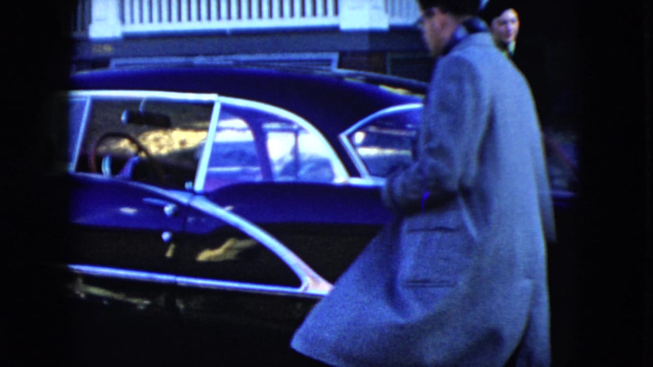 A man in a light overcoat approaches a dark blue sedan outside a Cleveland house in the 1951 home movie.