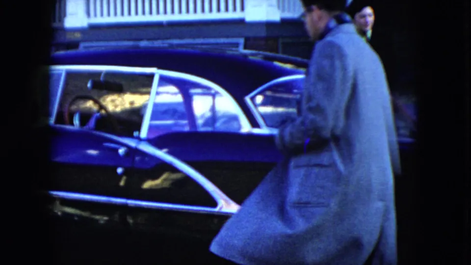 A man in a light overcoat approaches a dark blue sedan outside a Cleveland house in the 1951 home movie.