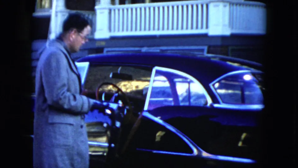 A man wearing gloves opens the driver's door of a dark blue sedan outside a house in 1951 Cleveland footage.