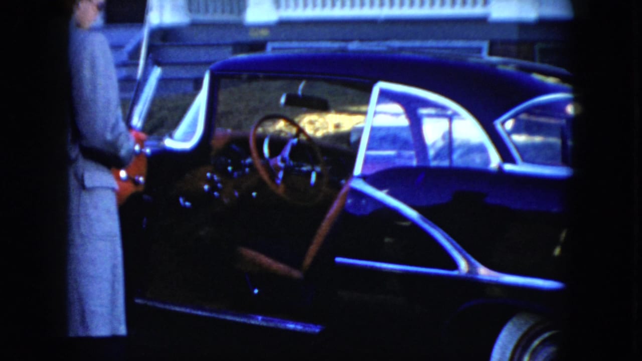 Man in a light suit stands beside an open vintage sedan door under nighttime light.
