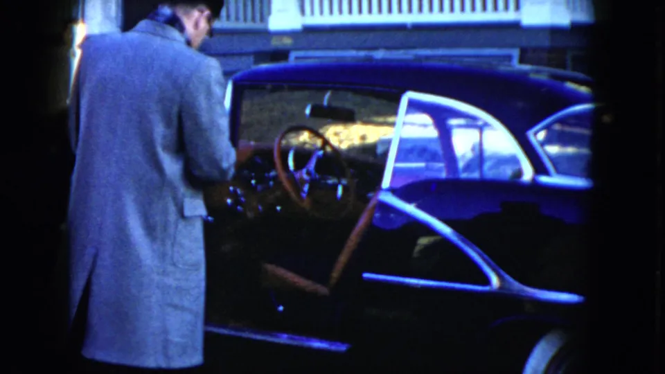 A rear view shows the man leaning toward the open door of the 1951 Cleveland sedan, with the dashboard and steering wheel partly lit.