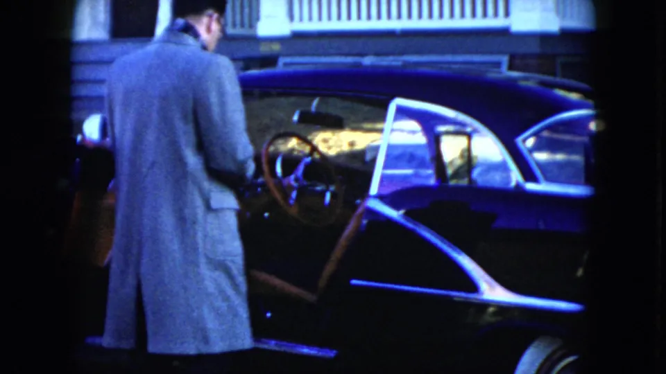 The 1951 Cleveland home-movie frame shows a man at the open sedan door, with the blue bodywork, chrome trim, and steering wheel visible.