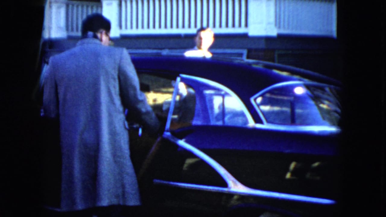 The man stands close to the open driver's door of a dark 1950s sedan in the Cleveland home-movie scene.