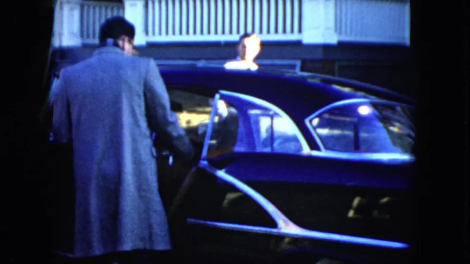 The man stands close to the open driver's door of a dark 1950s sedan in the Cleveland home-movie scene.