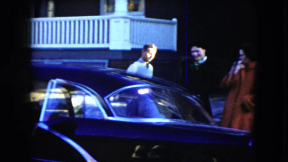 Pedestrians stand near a residential porch as the dark sedan fills the foreground of the Cleveland 1951 home-movie frame.