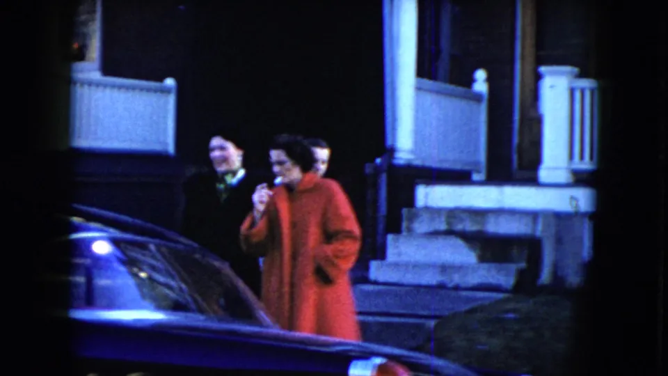 Two pedestrians, one in a red coat, move near porch steps while the dark sedan remains in the foreground in Cleveland, 1951.