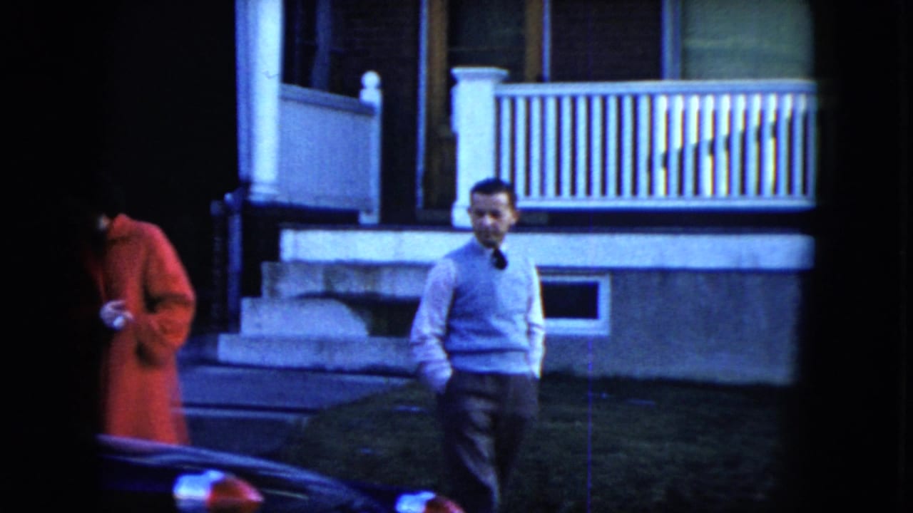 In 1951 Cleveland footage, a woman in a red coat and a man in a light sweater stand on a sidewalk outside a house with a white porch railing.