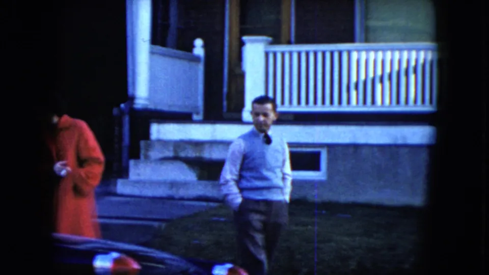 In 1951 Cleveland footage, a woman in a red coat and a man in a light sweater stand on a sidewalk outside a house with a white porch railing.
