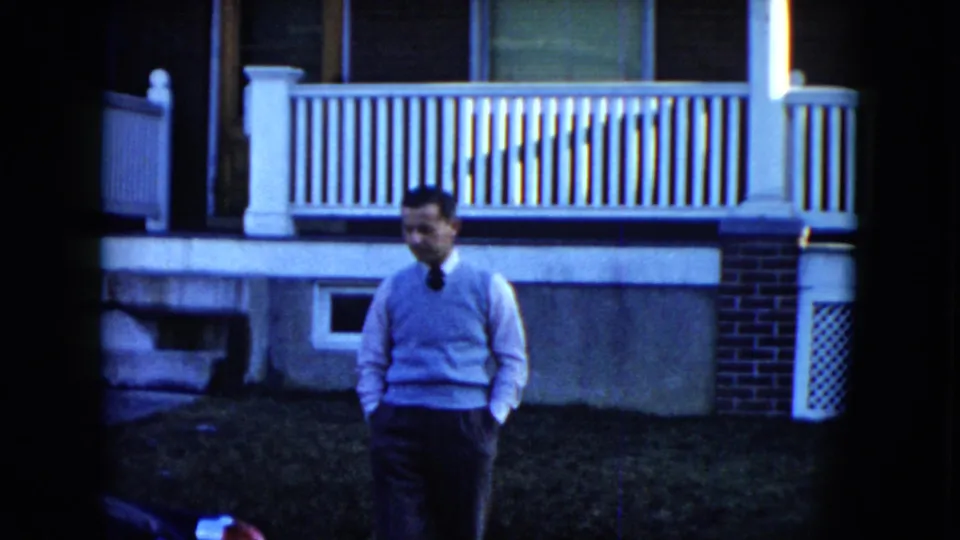 A man in a light sweater and dark tie walks toward the camera in 1951 Cleveland home-movie footage, with a porch and house behind him.