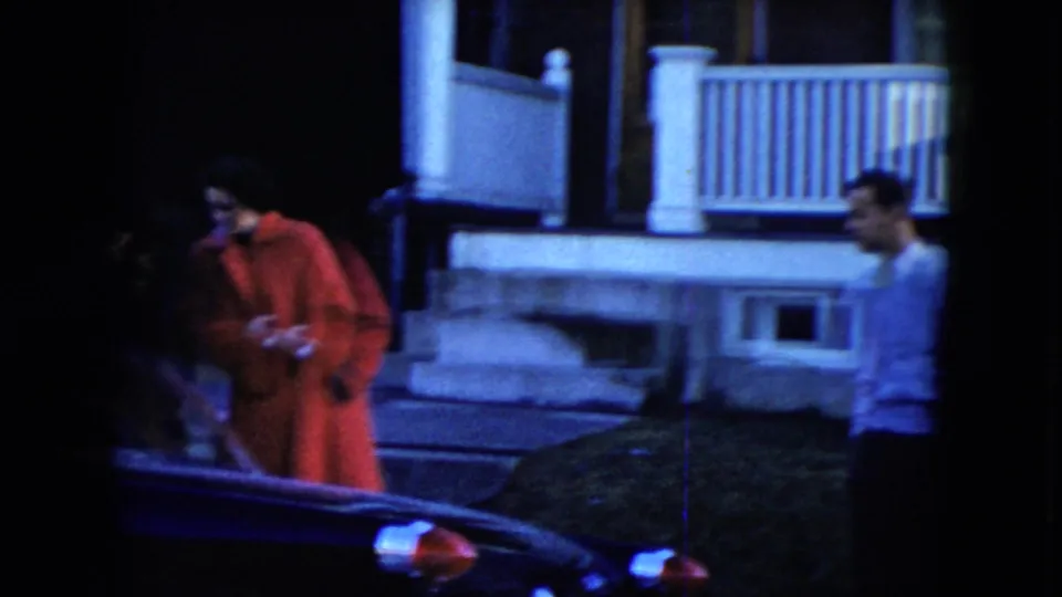 In a 1951 Cleveland exterior, a woman in a red coat stands near a parked car as a man in a light sweater moves at the right edge of the frame.
