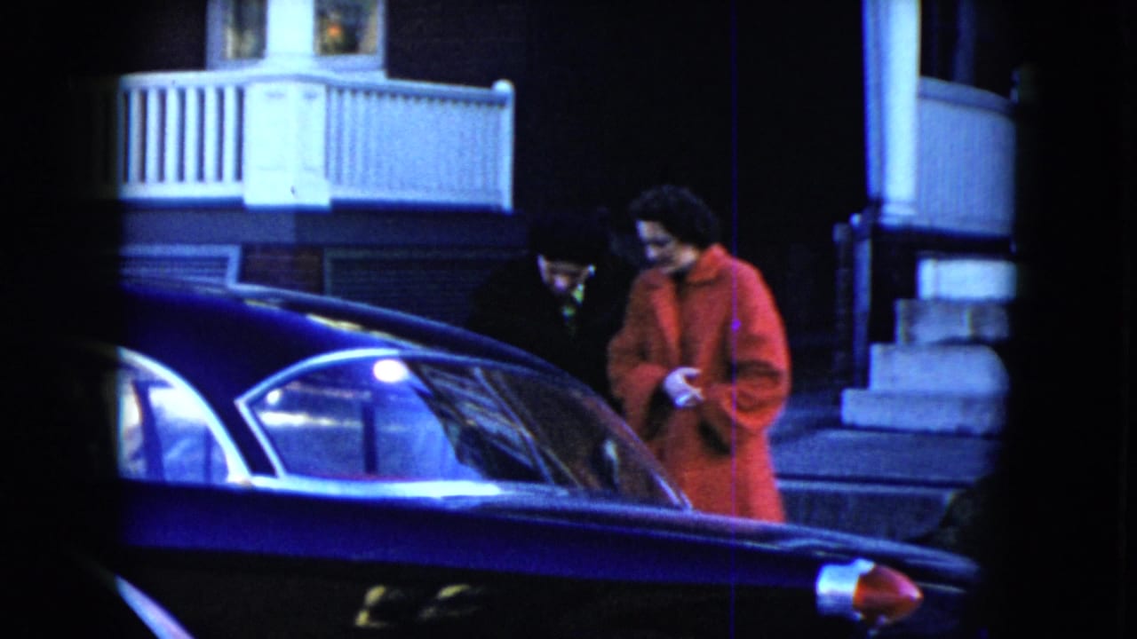 In the 1951 Cleveland home-movie scene, two adults lean toward a dark car, with porch railings and steps behind them.