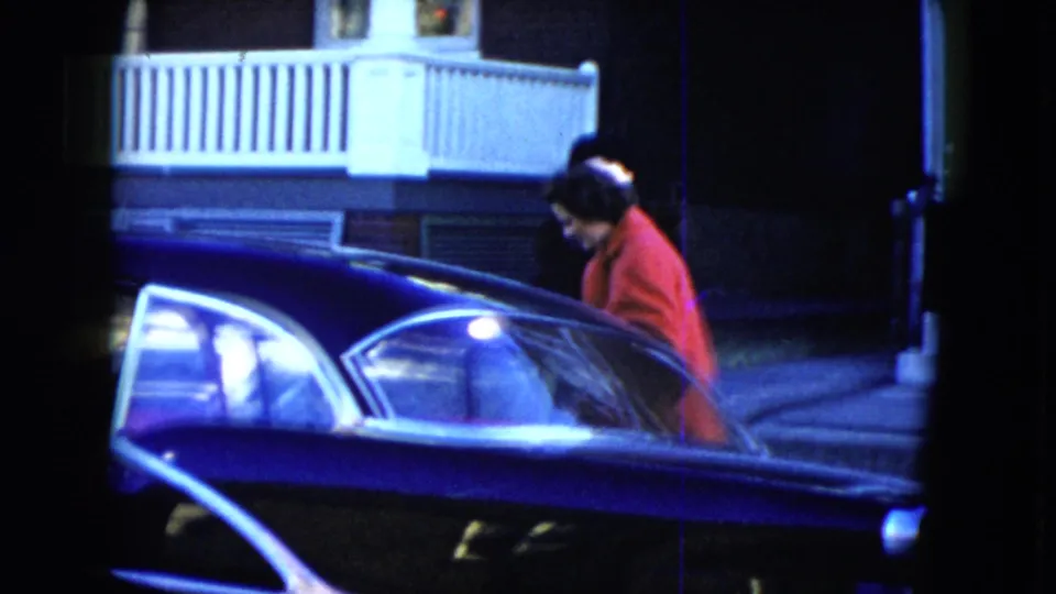 In Cleveland, Ohio, 1951, the woman in a red coat stands partly behind an open dark car door near a porch railing.