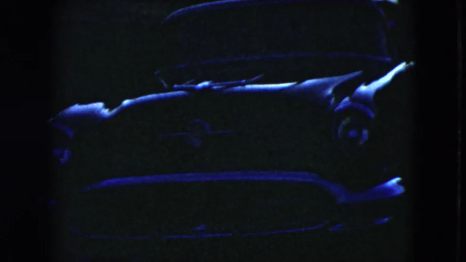 Grainy 8mm frame from the 1951 Cleveland clip showing a parked mid-century car with its windshield and hood faintly lit in blue.