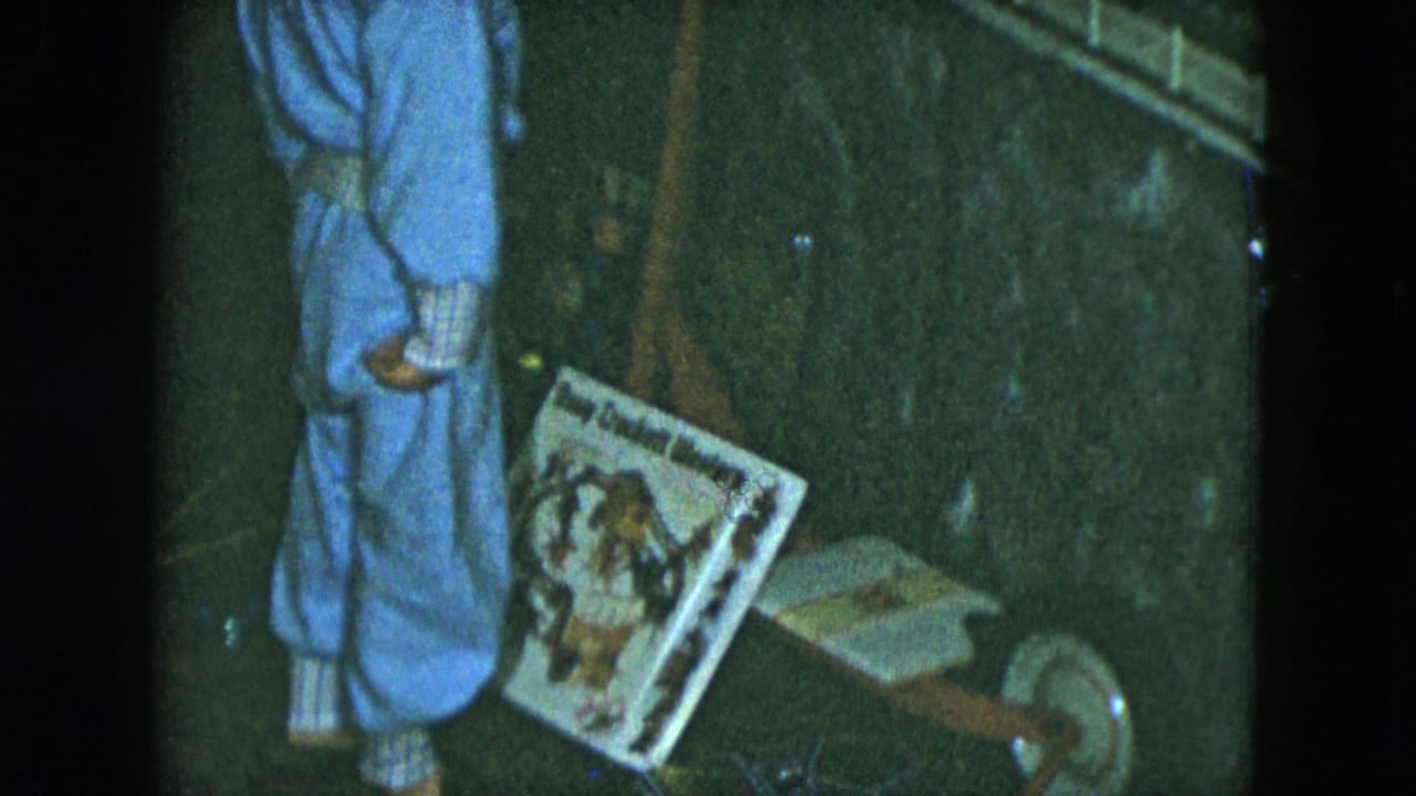 Boy in blue pajamas stands beside a boxed toy and small wheeled wooden toy on a dark floor.