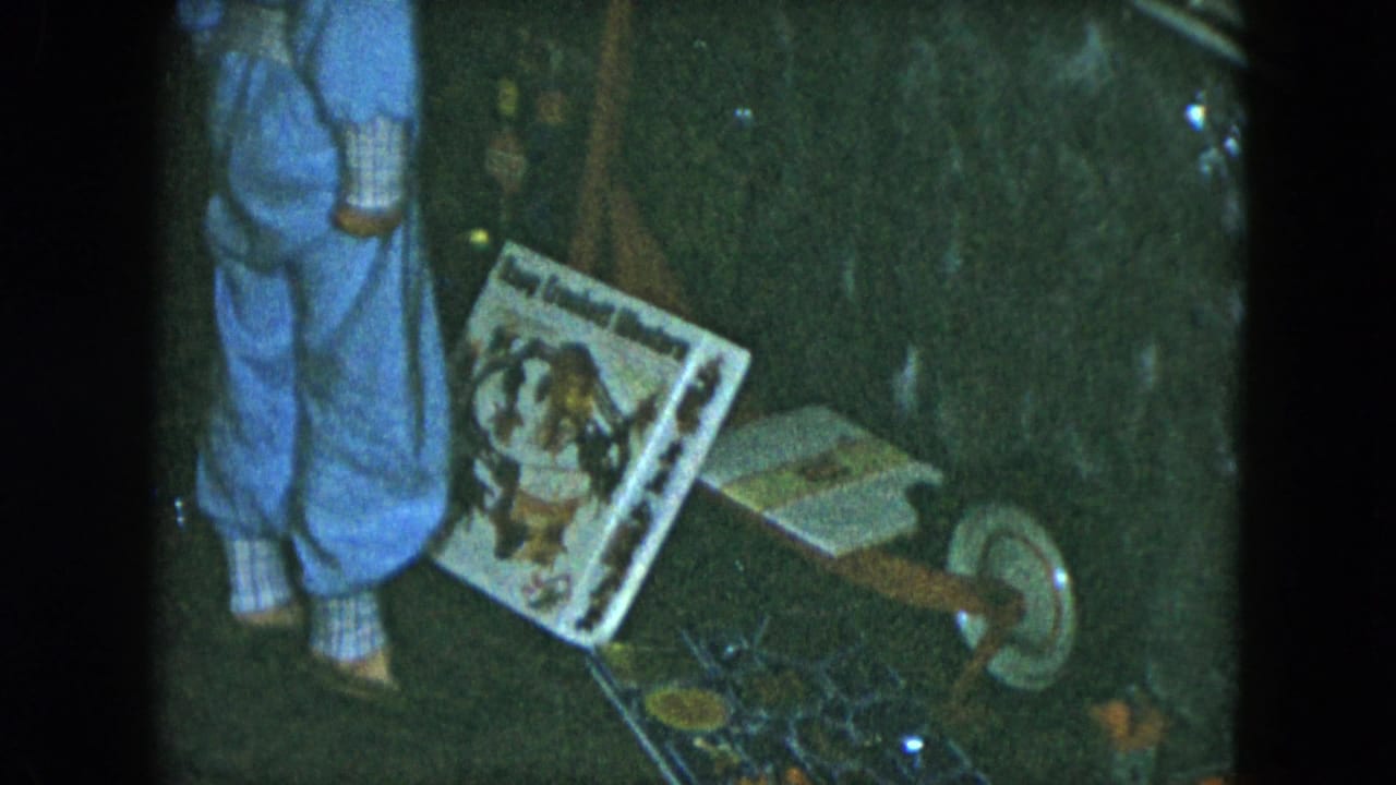 The boy remains partly in frame beside a boxed toy and wheeled wooden toy, with small play items scattered nearby in the dim interior.