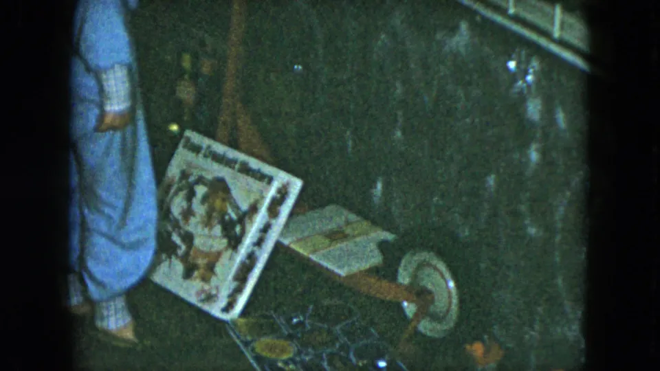 A closer 8mm frame shows the child's blue pajama legs next to a tilted toy package and a wooden wheeled toy in a dim room.