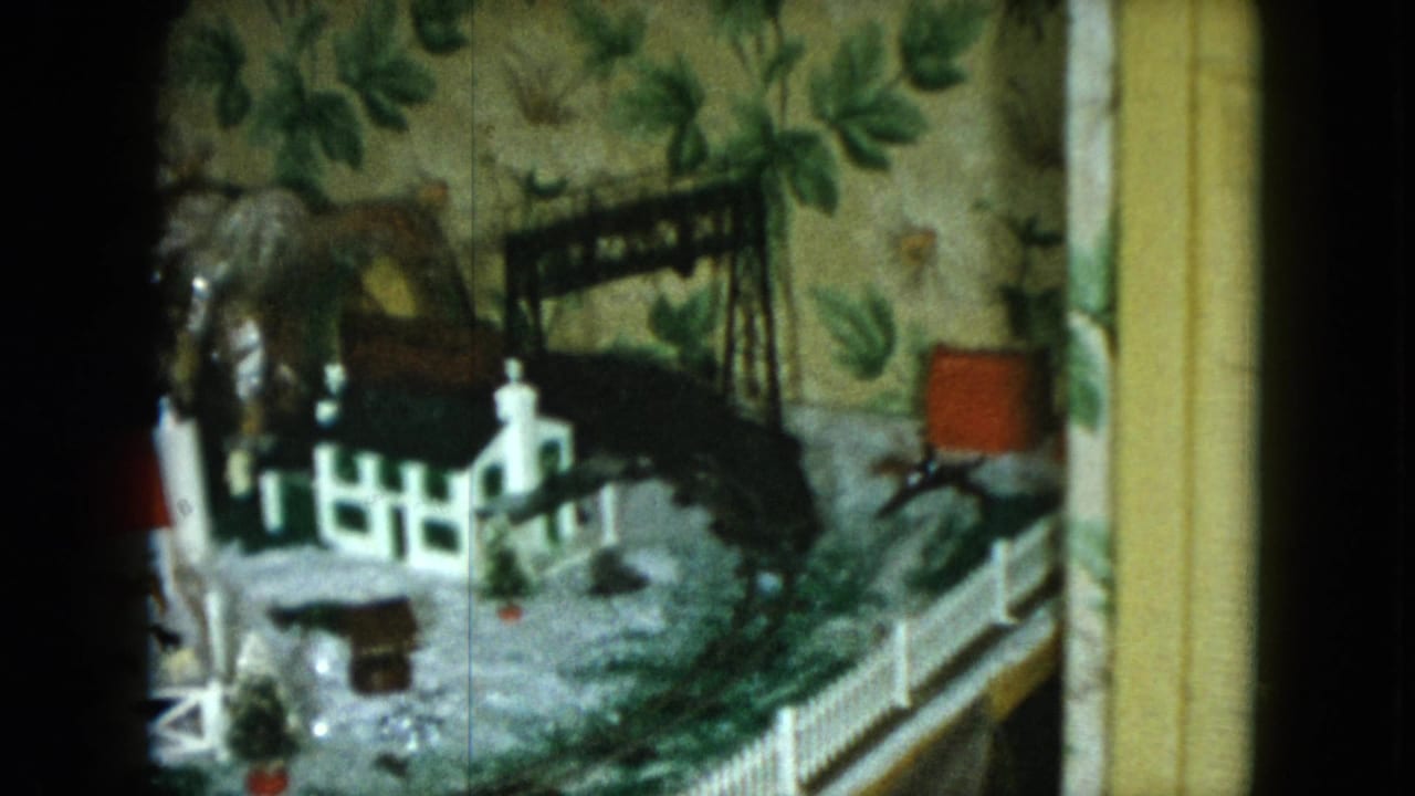 1951 Cleveland home-movie frame showing a small church-like building, bridge structure, fencing, and curved model train track on a tabletop.