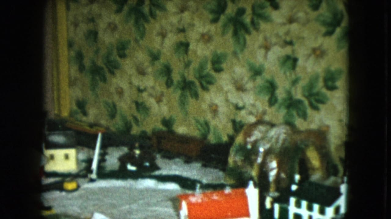 1951 Cleveland home-movie frame showing a tabletop model train set with small houses, rail track, and green floral wallpaper.