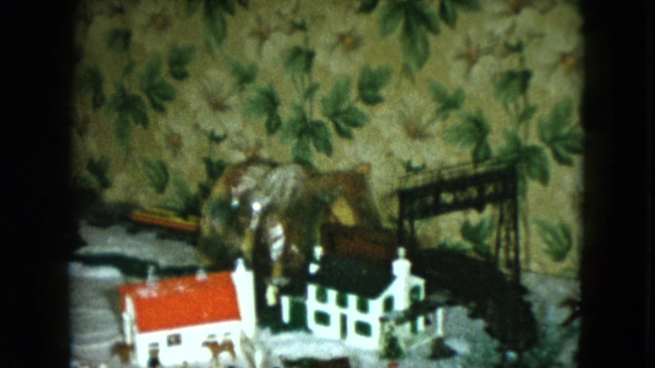 Miniature house and white fence beside a dark model train on a tabletop rail layout.