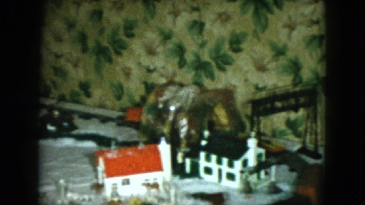1951 Cleveland home-movie frame showing a closer section of the model railroad with houses, fence, track, and patterned wallpaper.