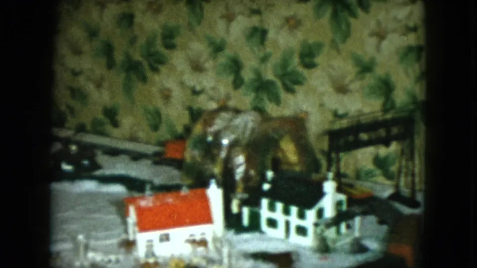 1951 Cleveland home-movie frame showing a closer section of the model railroad with houses, fence, track, and patterned wallpaper.