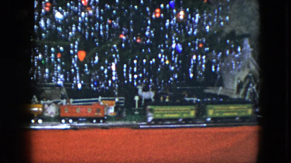 The 8mm frame shows model train cars moving past small houses, fences, and bright Christmas tree lights.