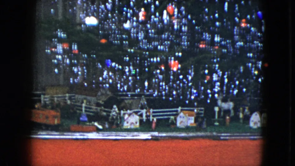 A wider view of the holiday train layout shows small houses, white fences, ornaments, and train cars under a lit tree.
