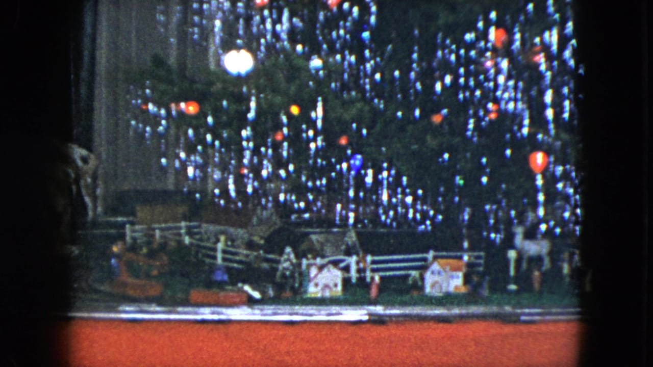 Model train display beneath a lit Christmas tree with miniature houses and a white fence.