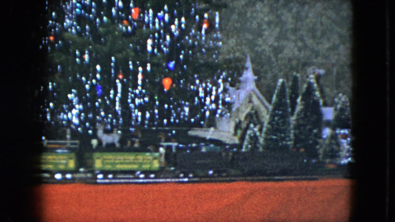Green and yellow model train cars move across a Christmas display with trees and a church.