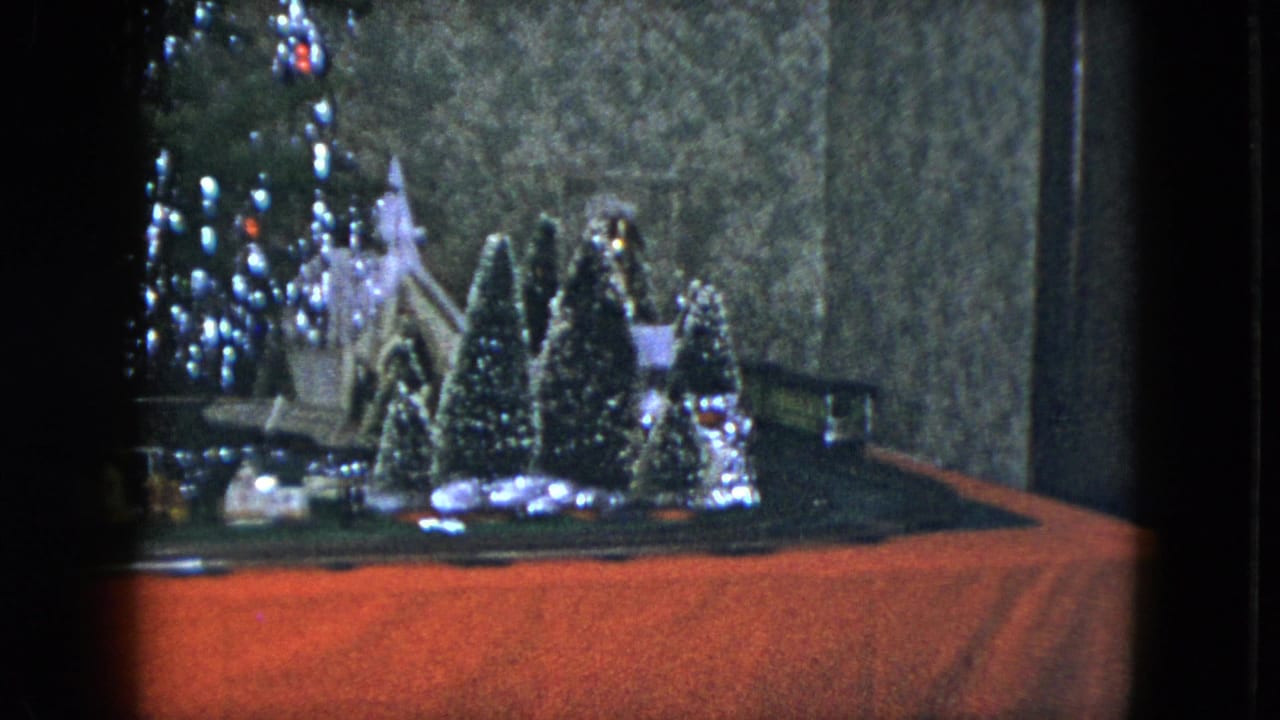 Model train track and miniature Christmas village beside a lit tree and red base cloth.