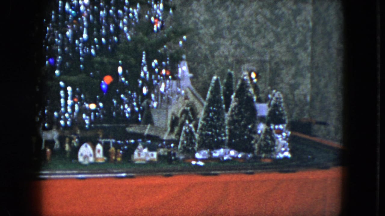 1951 Cleveland home-movie frame of a Christmas village with miniature houses, trees, train track, and glowing tinsel-lit tree.