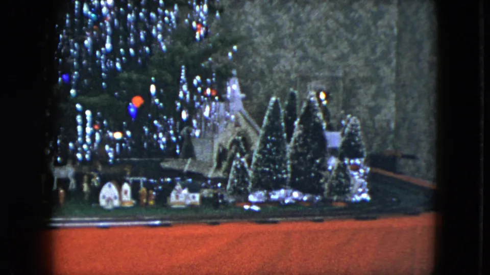 1951 Cleveland home-movie frame of a Christmas village with miniature houses, trees, train track, and glowing tinsel-lit tree.