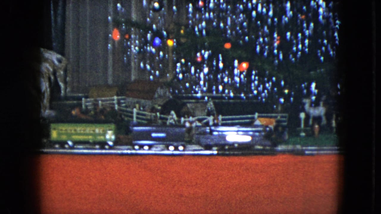 Model train cars pass beneath a lit Christmas tree beside a white fence and rocky tunnel.