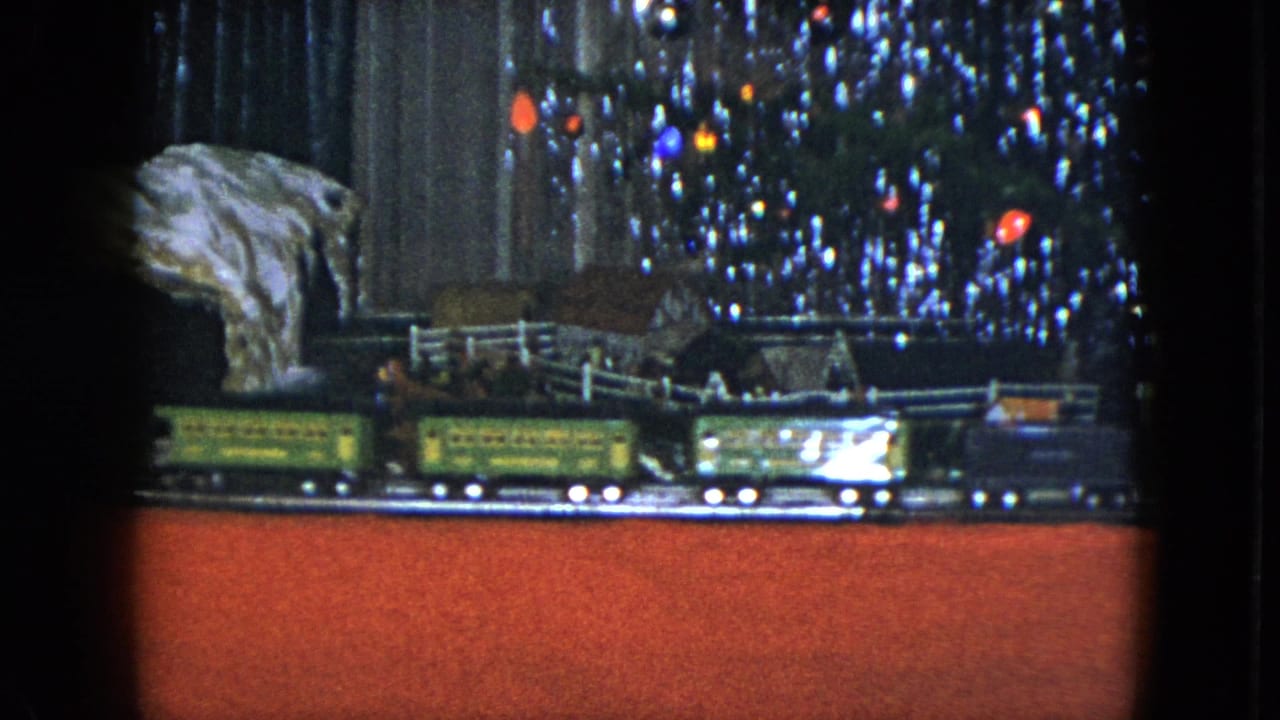 1951 Cleveland home-movie frame with green and dark model train cars moving below tinsel branches and warm tree lights.