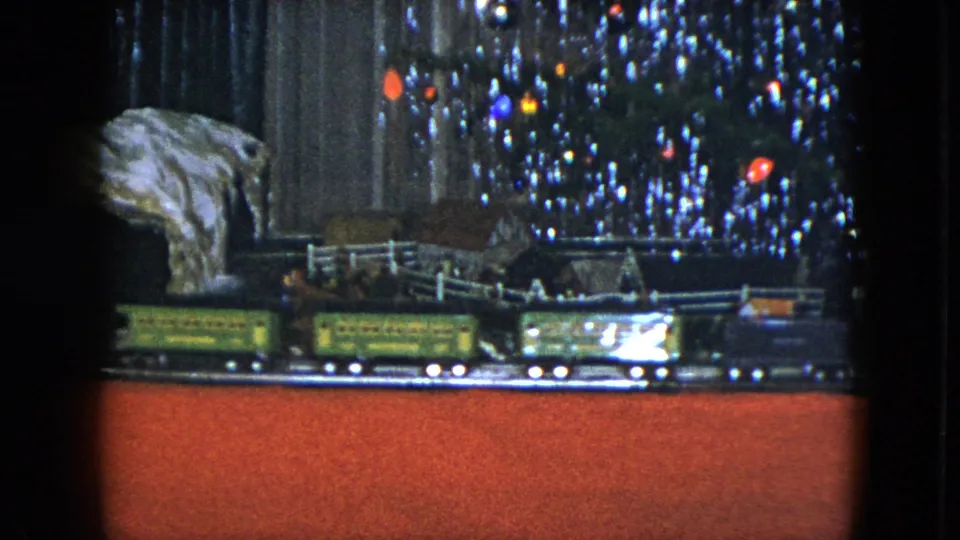 1951 Cleveland home-movie frame with green and dark model train cars moving below tinsel branches and warm tree lights.