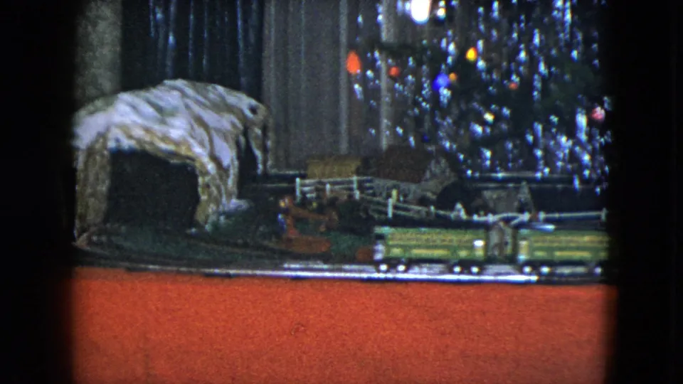 1951 Cleveland 8mm still showing the model railway layout with a rocky tunnel, white fence, train car, and decorated tree edge.