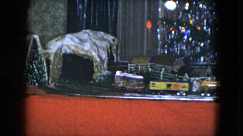1951 Cleveland home-movie frame showing model train cars beside a small tree, rocky tunnel, white fence, and lit Christmas tree branches.