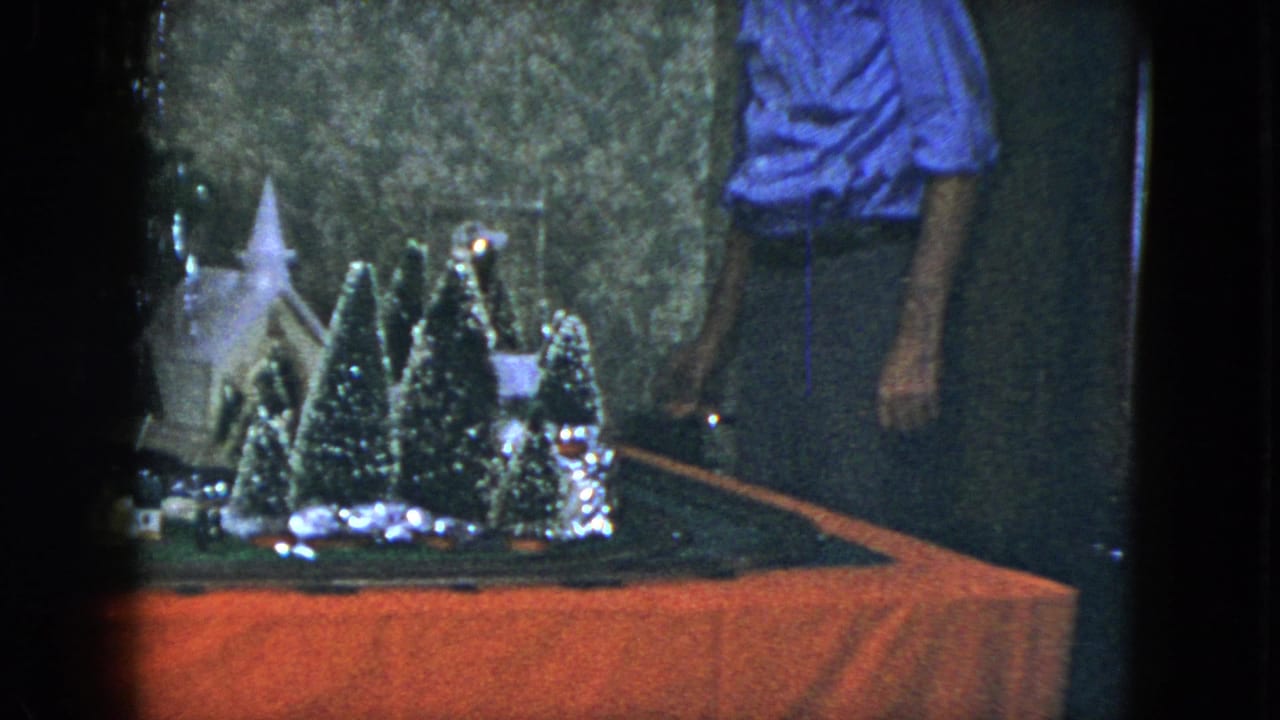 Tabletop holiday train layout with miniature trees, a small building and part of a man standing nearby.