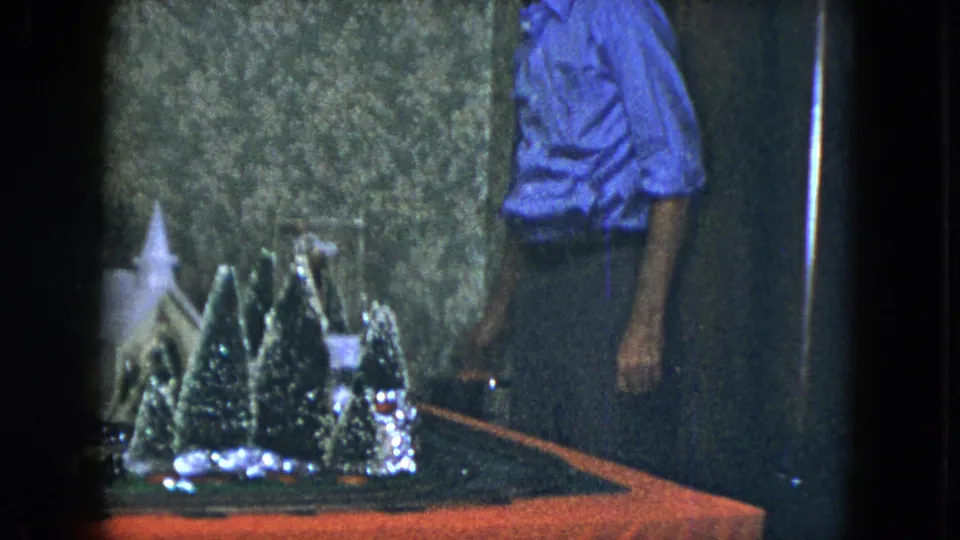 1951 Cleveland home-movie frame showing an adult man next to a model train holiday display with snowy trees and a small church-like structure.