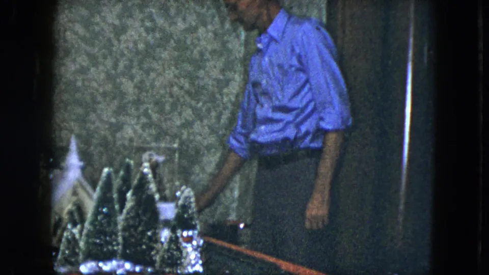 1951 Cleveland frame of an adult man reaching toward the tabletop Christmas train set, with miniature evergreens and a small building visible.