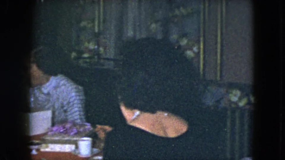 The handheld 8mm frame shows two adult women turned partly away from the camera in a dark dining room setting.