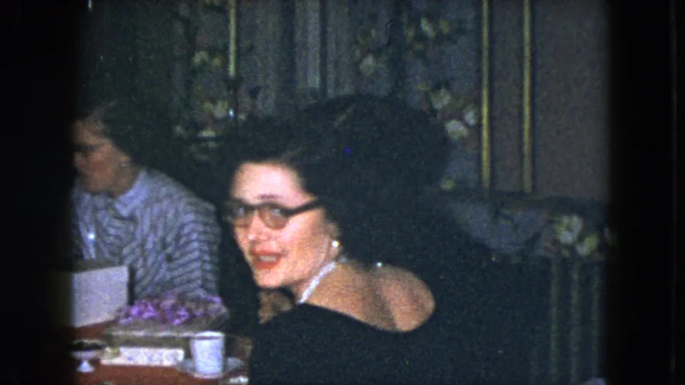 In the Cleveland 1951 dinner scene, a woman with glasses turns toward the camera while another woman remains seated behind her.