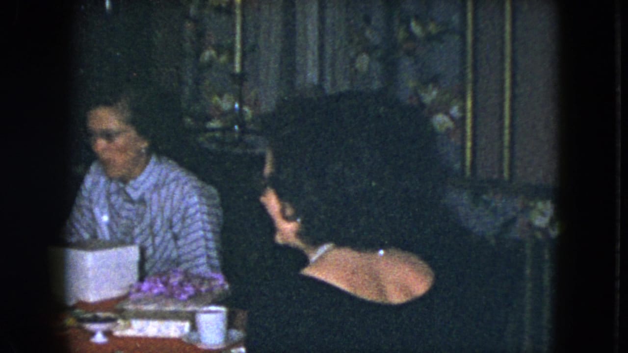 1951 Cleveland home-movie frame of two women at a red dining table with candles, food, flowers, and a white box nearby.