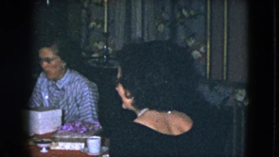 1951 Cleveland home-movie frame of two women at a red dining table with candles, food, flowers, and a white box nearby.